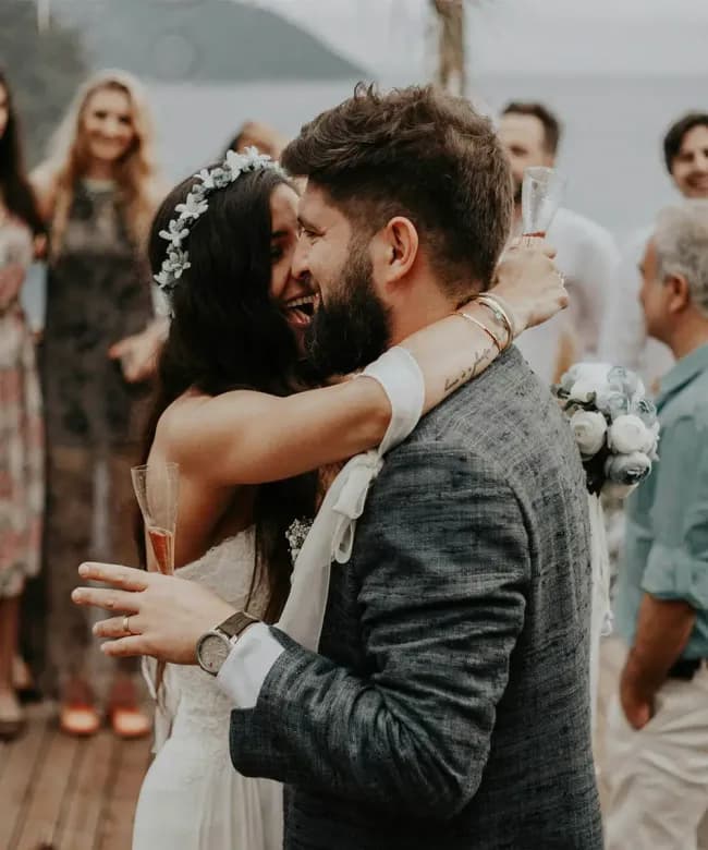 A close-up of a happy bride with a flower crown and a groom in a grey linen suit hugging and laughing. They are holding champagne glasses at an outdoor event with blurred guests in the background.