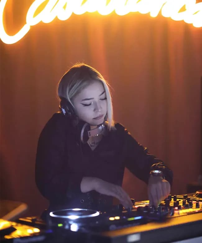 A blonde female DJ wearing headphones and a black outfit mixing music on a professional controller. A warm yellow neon sign glows in the background of the dimly lit venue.