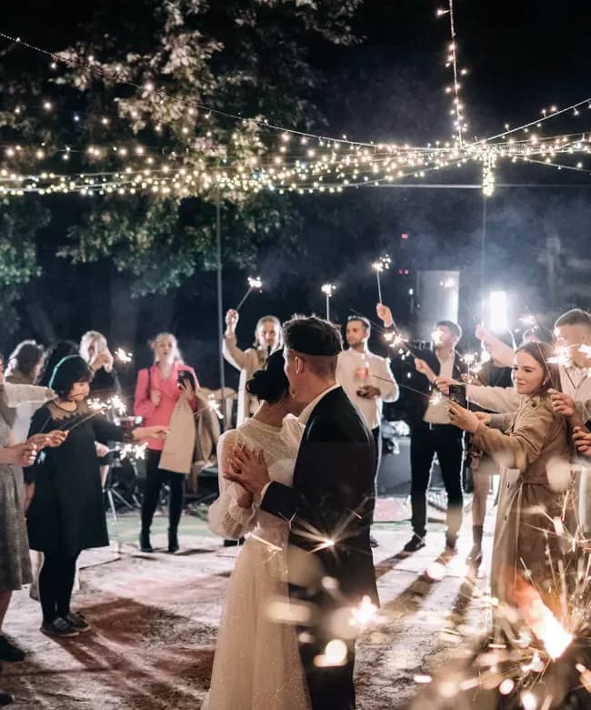 A bride and groom sharing their first dance at an outdoor evening wedding reception. Guests surround them holding glowing sparklers under a canopy of decorative string lights.