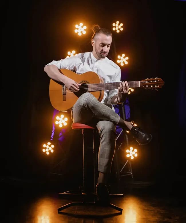 A male musician with a bun playing a classical acoustic guitar while sitting on a stool. Warm stage lighting with circular vintage-style bulbs creates a bokeh effect in the background.