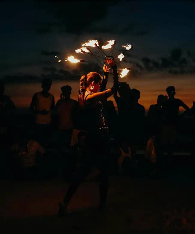 A female fire performer dancing with a lighted fire fan at night. She is silhouetted against a dark sky and a crowd of spectators watching the entertainment.