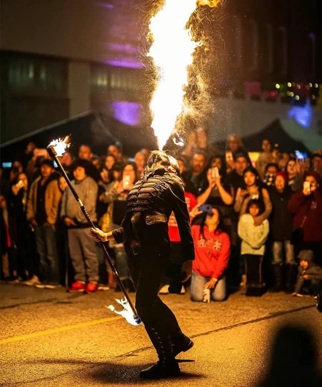 A professional fire breather exhaling a large plume of fire into the air during a night performance. An audience stands in the background watching the street entertainment.
