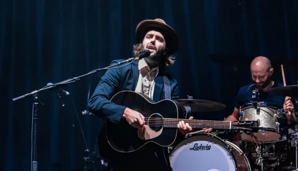 A male singer-songwriter wearing a hat and suit playing an acoustic guitar and singing into a microphone on stage with a drummer in the background.
