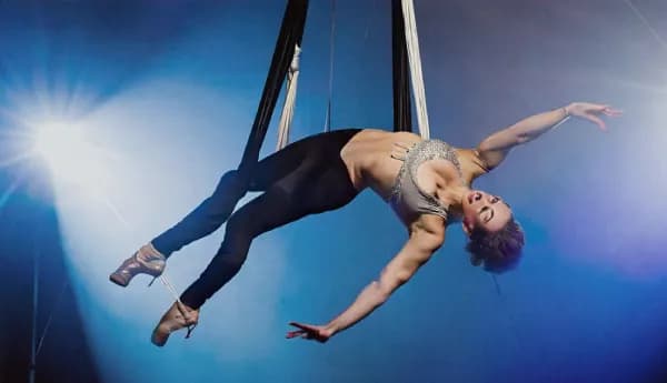 A female aerialist performing an acrobatic routine on black and white silks. She is arched backward in mid-air against a blue-lit stage background.