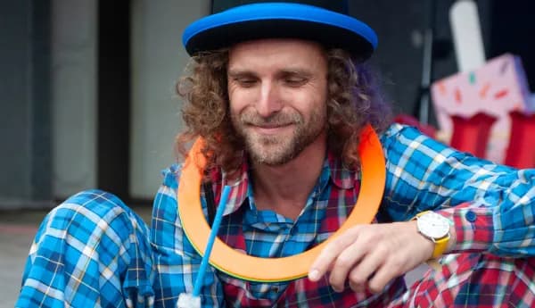 A male children's entertainer with curly hair wearing a colorful plaid shirt, a hat with blue trim, and an orange ring around his neck while performing for kids.