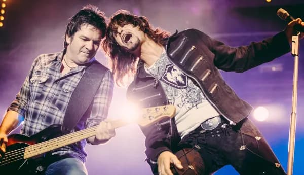 A high-energy live performance of a rock band featuring a bassist in a plaid shirt and a lead singer with long hair in a black jacket singing into a microphone.