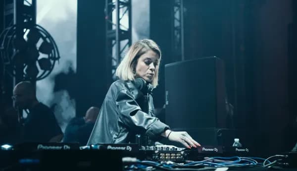 A female DJ with blonde hair wearing a black leather jacket, focused on mixing music on a professional controller at a concert or nightclub.