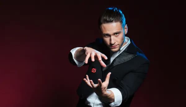 A male magician in a black suit gesturing with his hands as a red die levitates between them against a dark red background.
