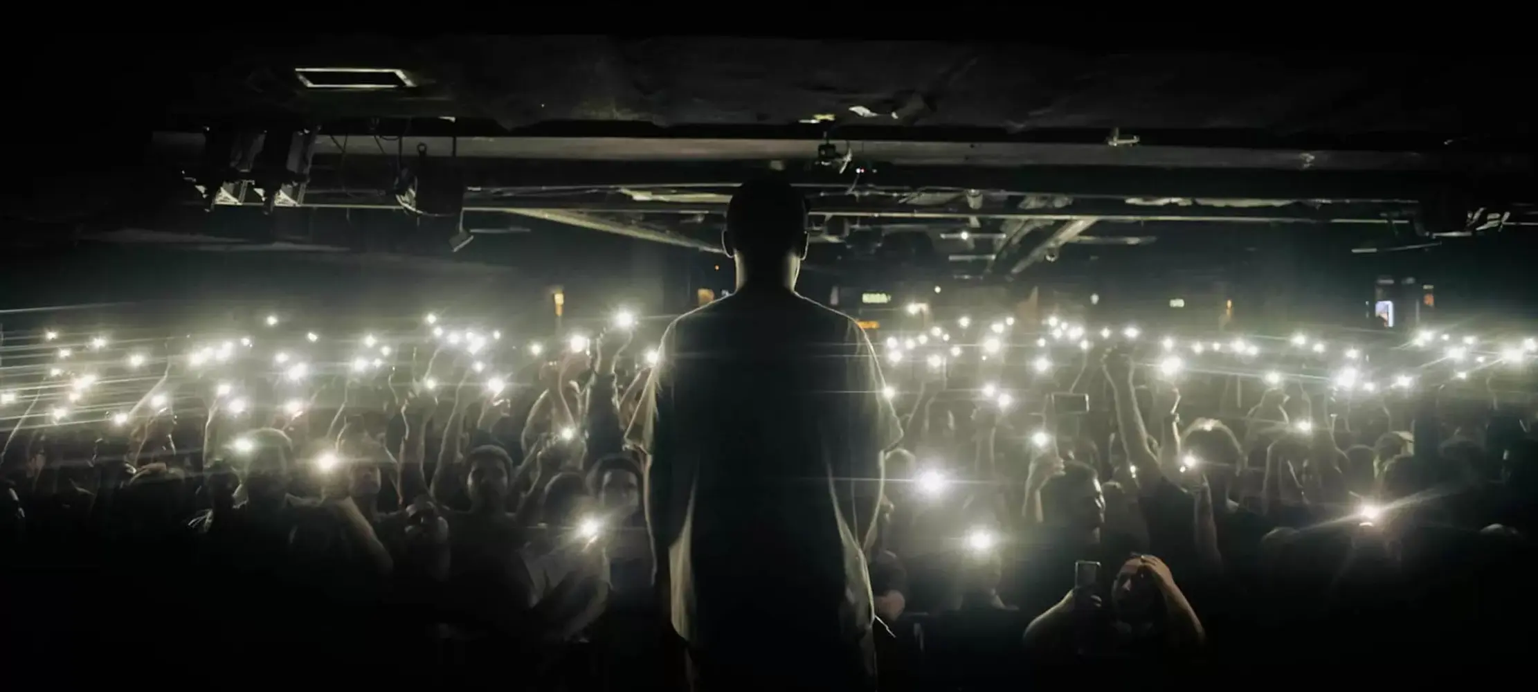 Silhouette of a professional live performer facing an engaged crowd with glowing phone lights in a dark venue, illustrating high-energy event atmosphere.